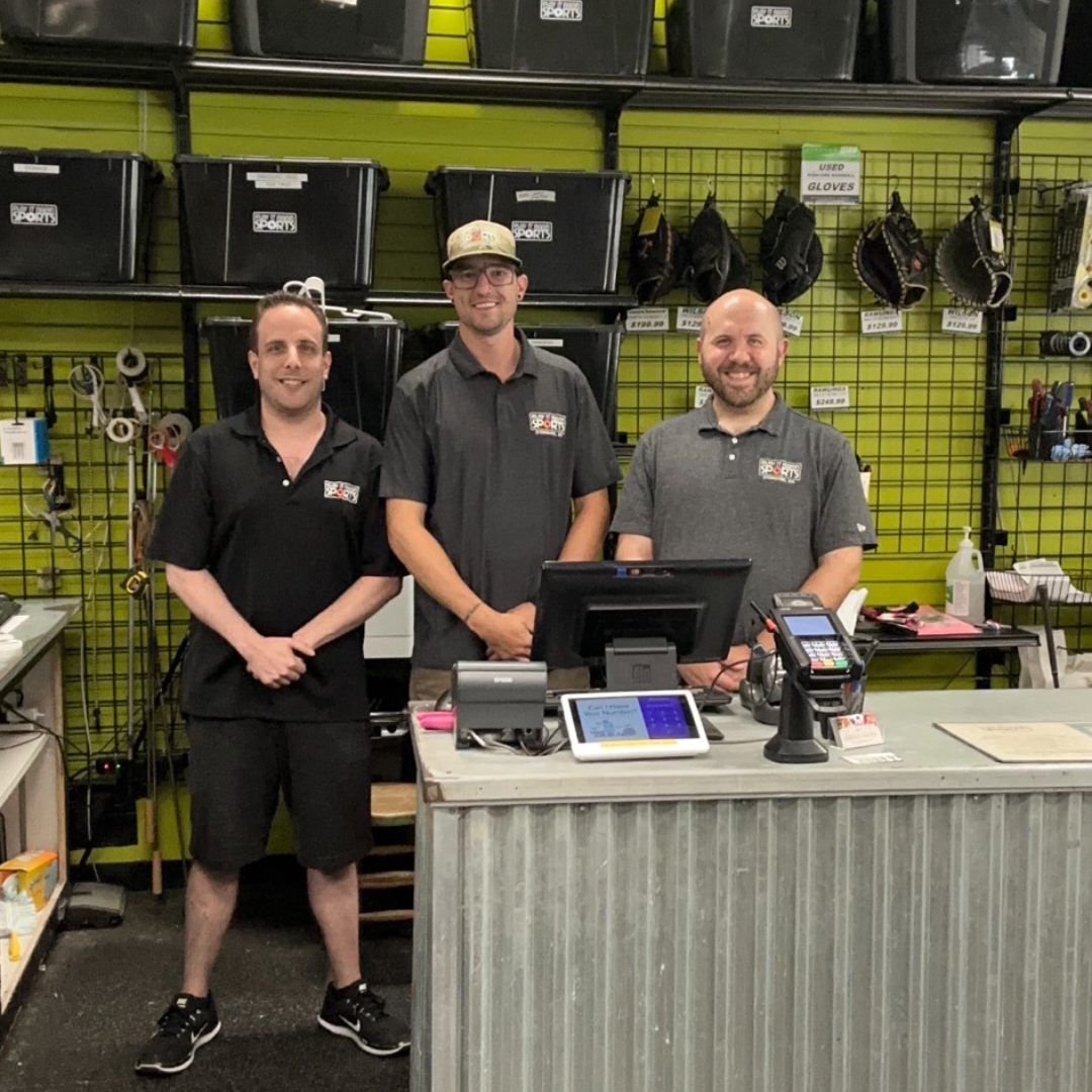 three men stand behind a counter, smiling at customers at Play It Again Sports Stoneham
